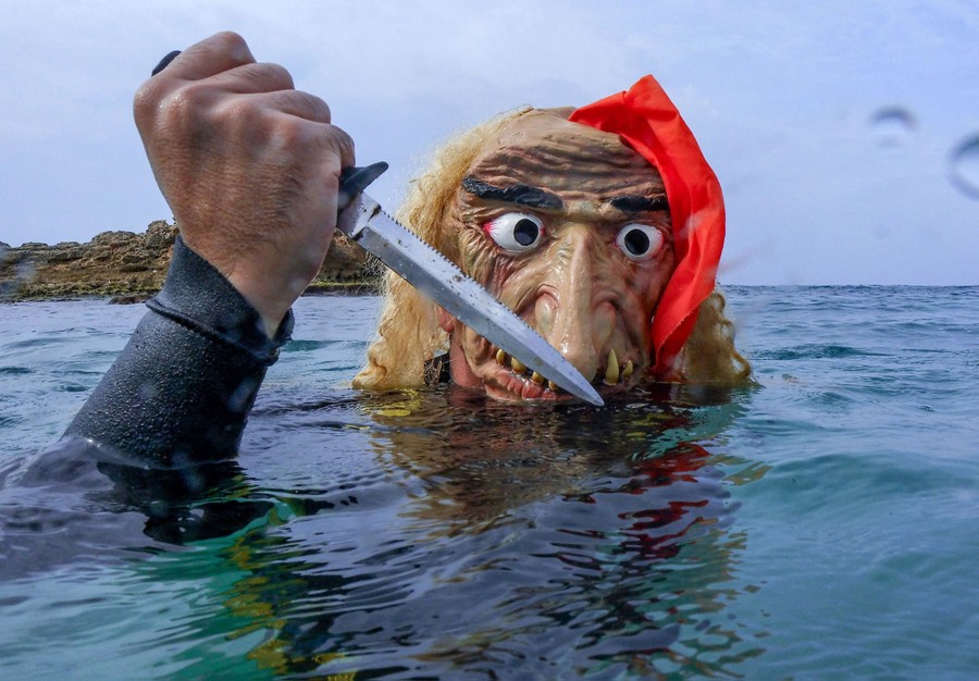 A person wearing a Halloween mask swims while holding a small knife.