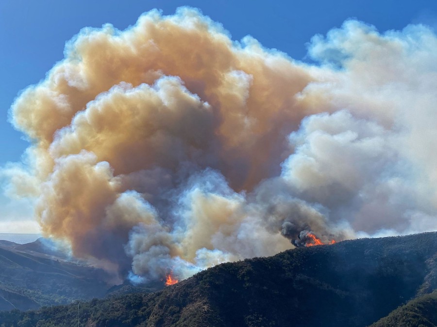 A large plume of smoke rises above burning hillsides.