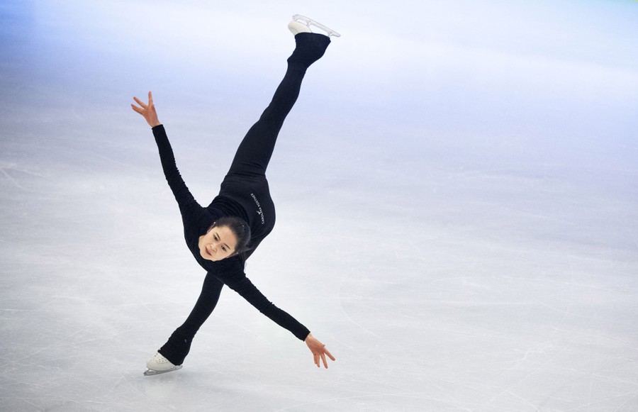 An ice-skater practices on the ice, skating on one foot, with the other foot raised high behind her.