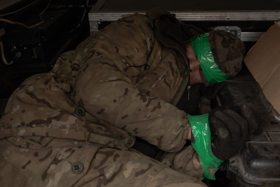 A prisoner of war, with tape wrapped over their eyes and around their wrists, lies in the back of a vehicle.
