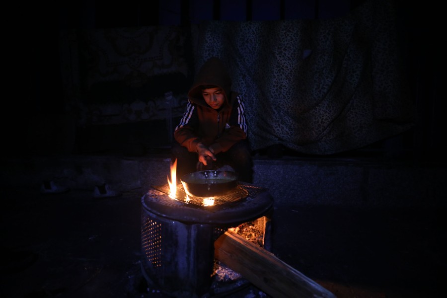A young person sits beside a wood fire, holding a pan on a grill over the flames.