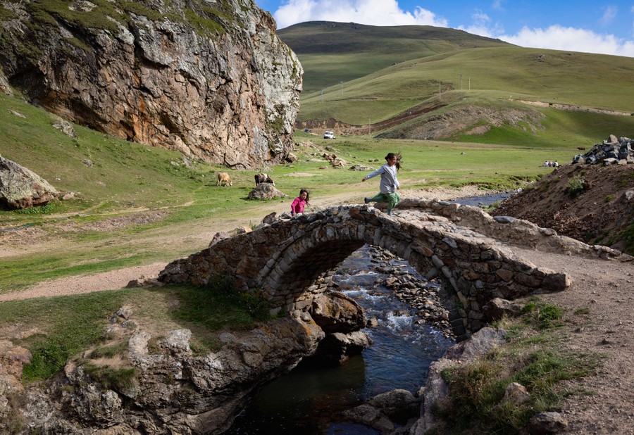Children play on a small stone bridge over a stream near grassy, treeless hills.