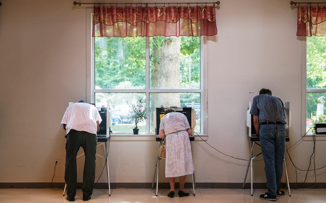 Three people filling out ballots in front of a window; trees visible through the window