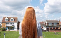 A white girl with long red hair, wearing a gray t-shirt, has her back to the camera and looks out at two lawns and two houses on a suburban street.