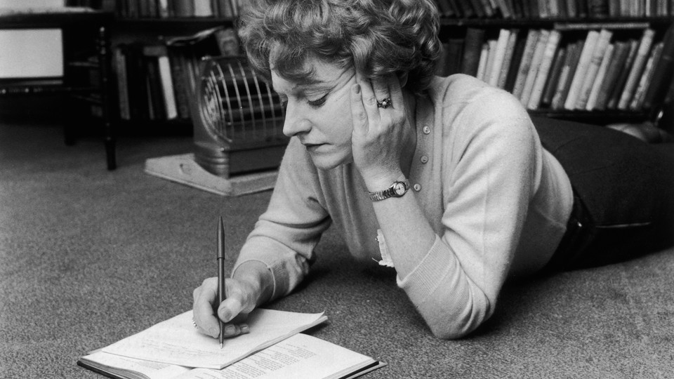 A photograph of Muriel Spark writing on a notebook on the floor