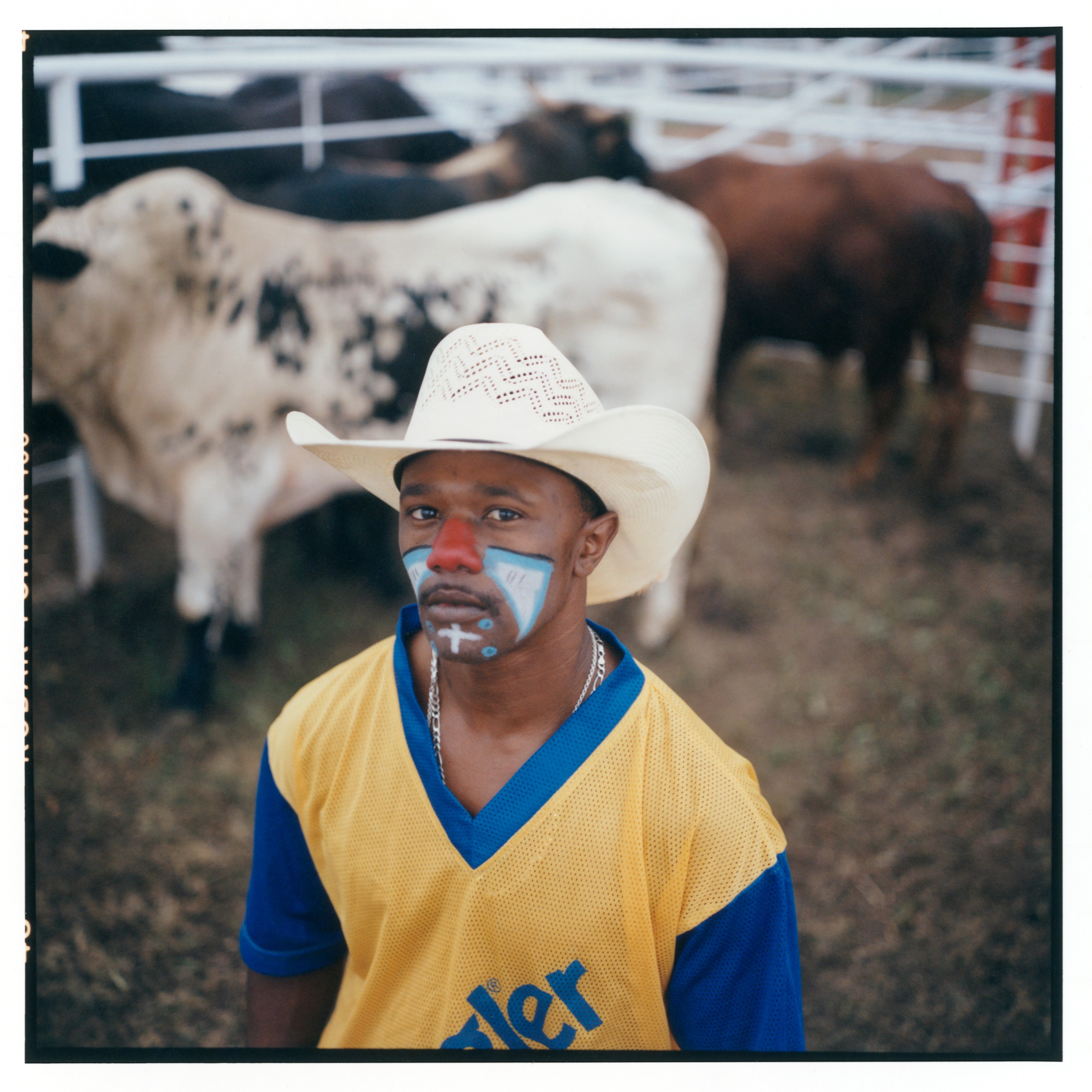 photo of Black man in yellow/blue Wrangler tee and straw cowboy hat with rodeo clown makeup on face, with bulls and arena in background