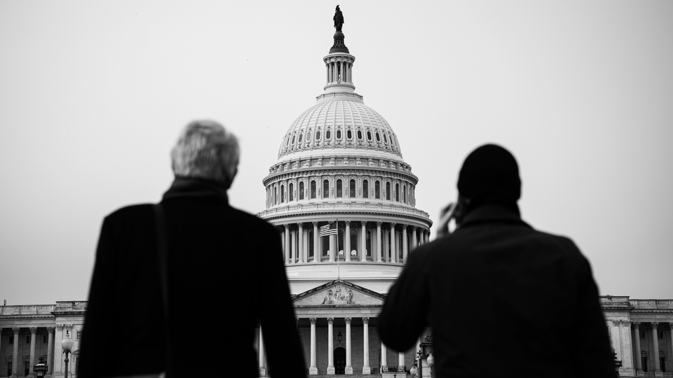 Black and white photograph of two men looking up at the US Capitol building, one holding a phone to his ear