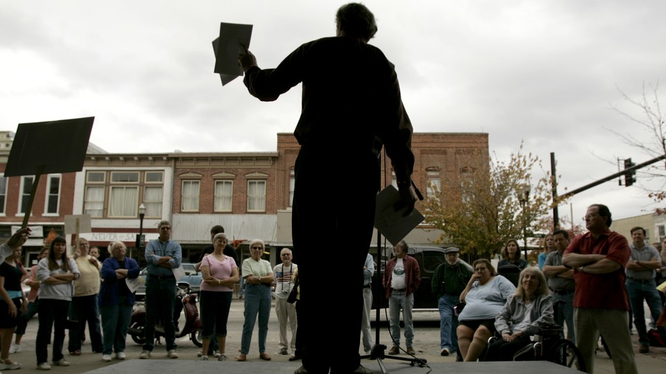 Al Norman, seen in silhouette, holds flyers and speaks to a crowd from a stage.