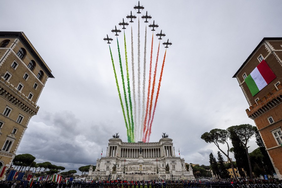 Nine aerobatic aircraft fly in a triangle formation above a piazza, trailing colored smoke.
