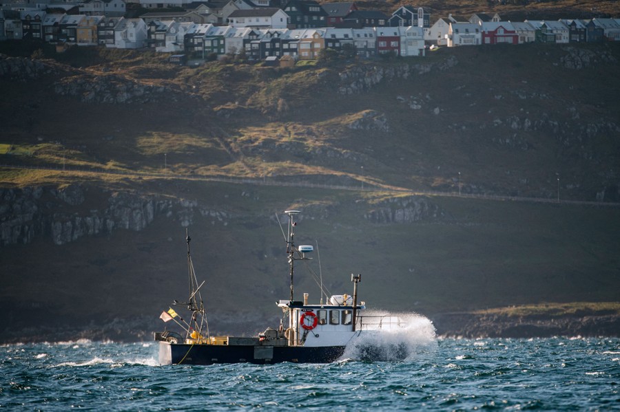 A fishing boat passes near an island. A row of houses can be seen on a hill above the shore.
