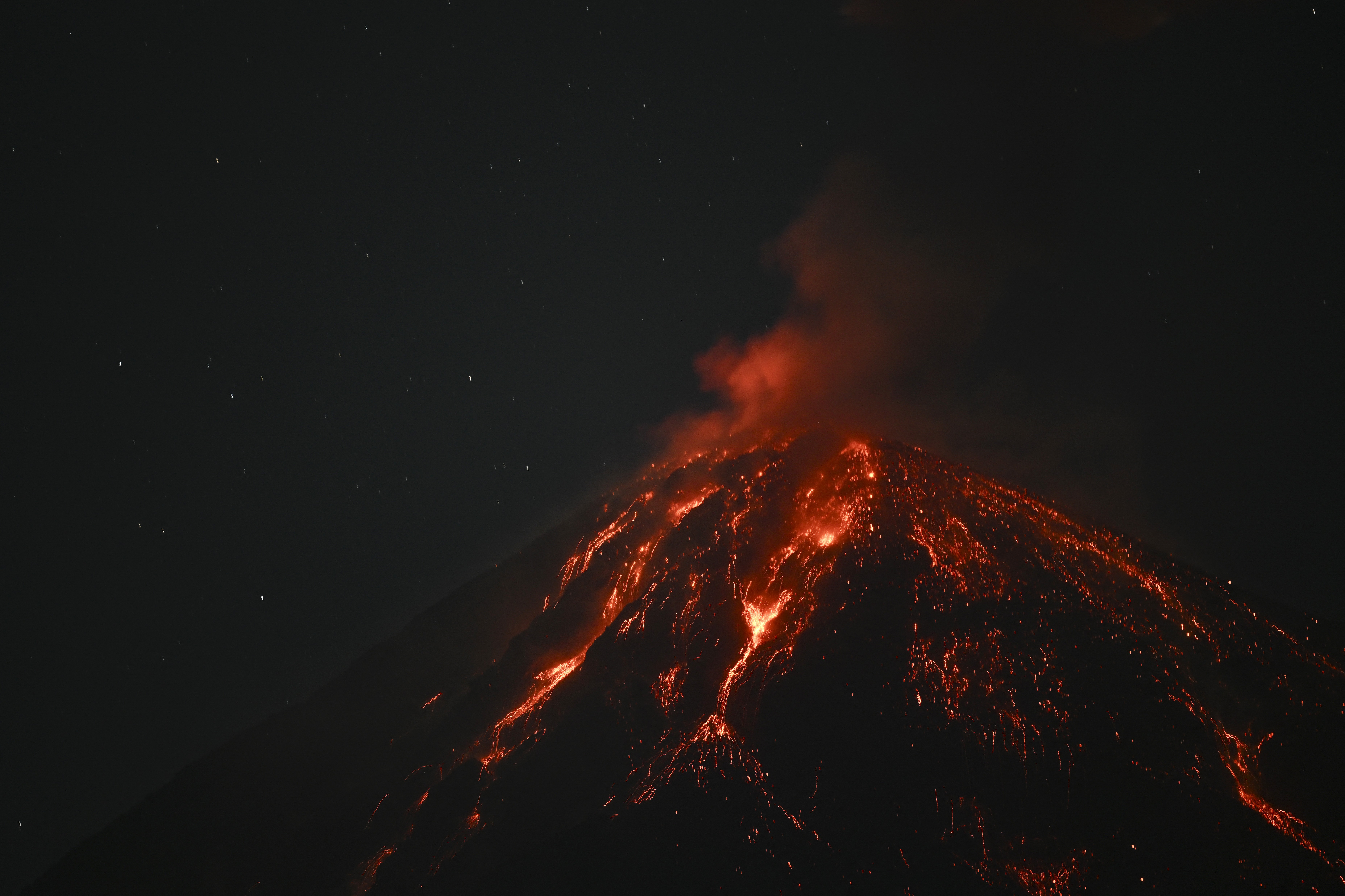 Lava glows across the top of an erupting volcano, seen at night.
