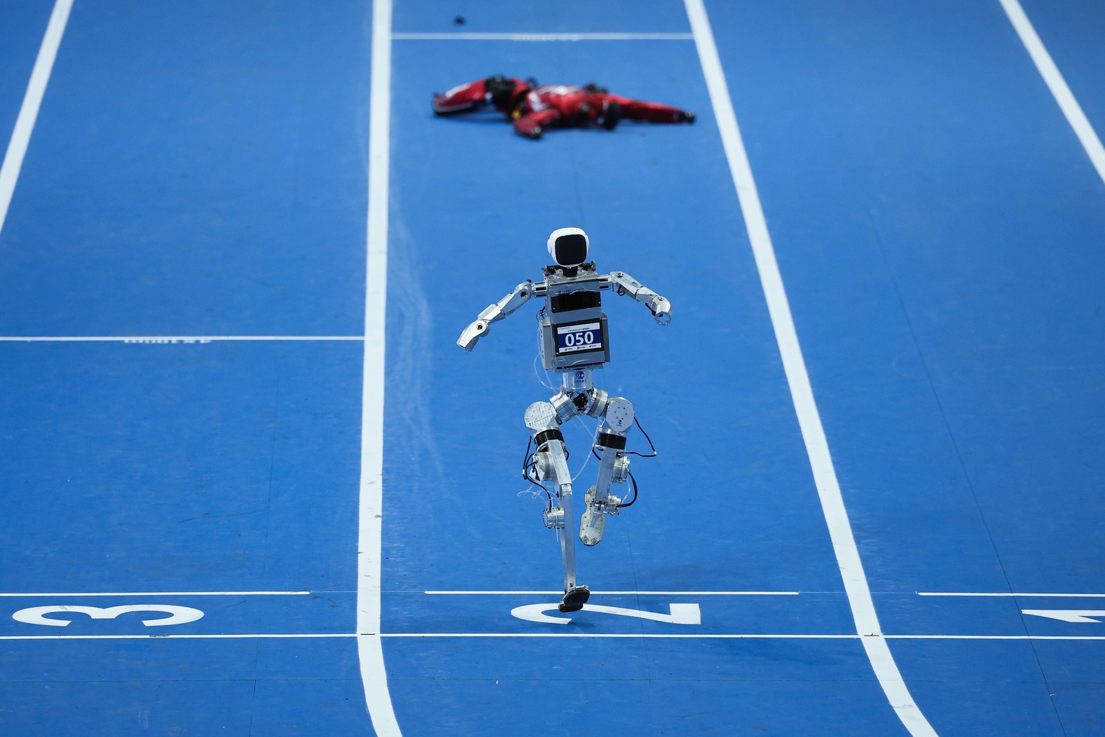 A humanoid robot crosses a finish line during a race. Another robot lies prone in the background.