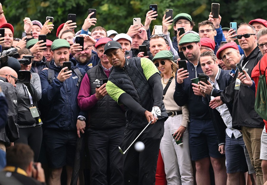 A crowd of onlookers—most holding up mobile phones—watch as Tiger Woods hits a golf ball.