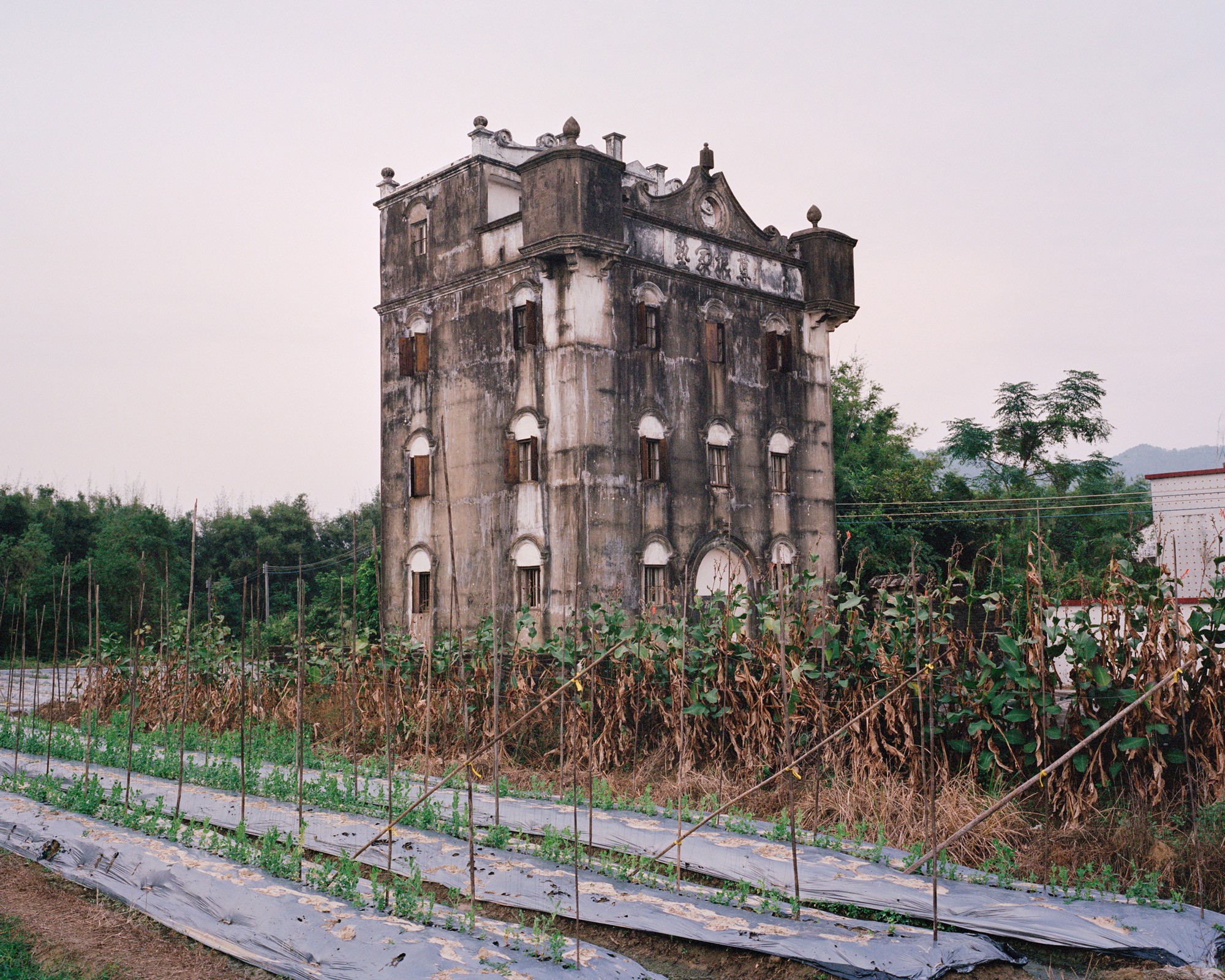 A weathered four-story watchtower stands above a field and smaller buildings.