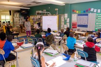 First-grade students are seated in a classroom listening to their teacher.