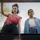 A still image of Margaret Qualley and Geraldine Viswanathan in “Drive-Away Dolls,” wearing puzzled expressions as they look into the trunk of a car. A gray-blue sky and green treetop are behind them.