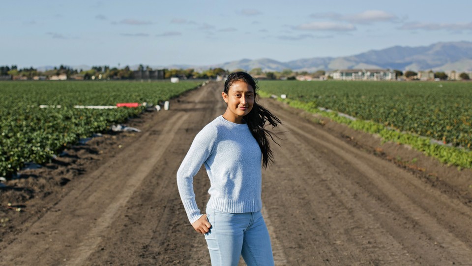 A young girl wearing a blue sweater and jeans stands on a dirt path in a field