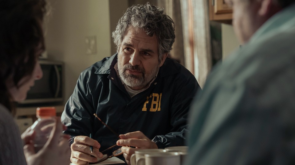 A color still from the show "Task" showing Mark Ruffalo wearing an FBI jacket and looking at a man and a woman in the foreground.