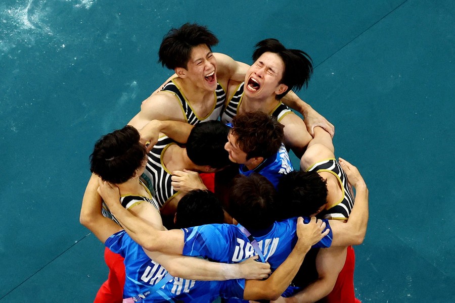 A group of gymnasts and coaches gather in a group hug, shouting and celebrating.