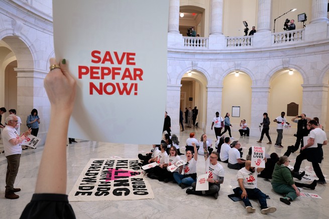 Someone holds a sign that says "save PEPFAR now" in a building with other protestors