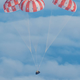 A gumdrop-shaped spacecraft, charred from the heat of reentry, coasts through blue sky beneath a trio of red-and-white parachutes.