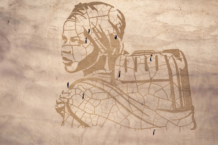 A large portrait carved into beach sand, and seen from above, depicts a child carrying a water container.