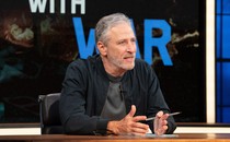 Jon Stewart leaning against a desk, holding a pen