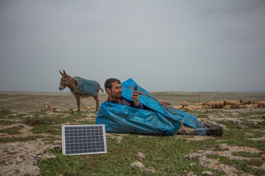 A shepherd lies near a small solar panel as he plays with his phone.
