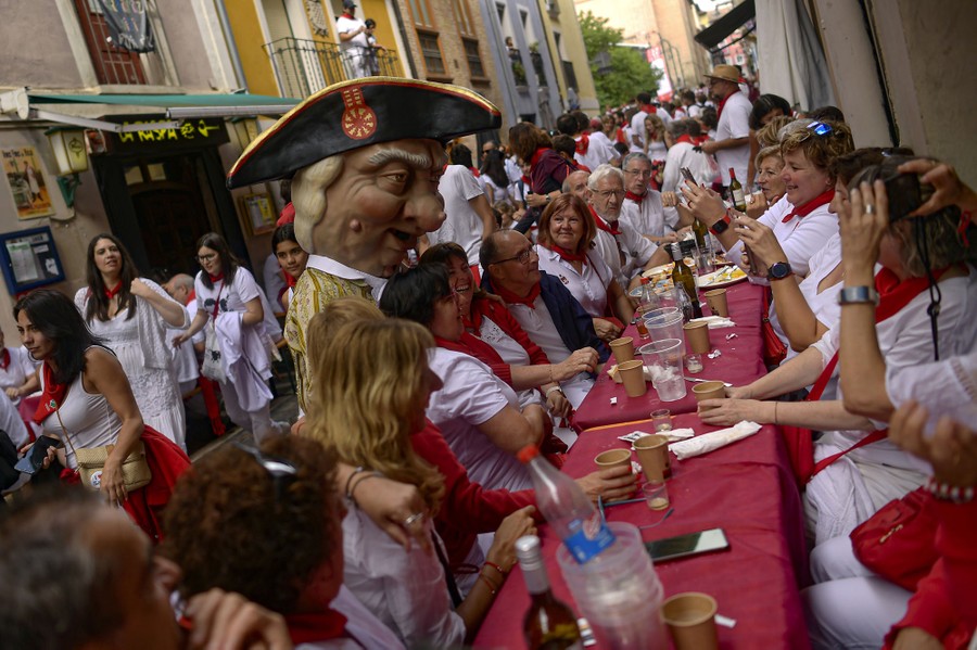 A person wearing a costume with a large head poses for photographs alongside people eating lunch at a long table, set up in a narrow street.