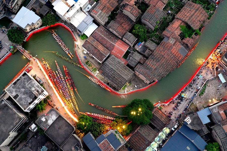 An aerial view of more than a dozen dragon boats in a twisting river surrounded by houses