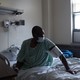 A masked man in a green gown sits up in a hospital bed, wearing medical cuff around his upper arm