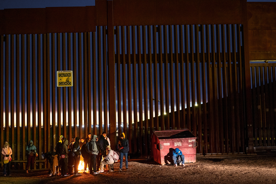 photo of group of people standing next to border wall with "Caution/Cuidado" sign and dumpster