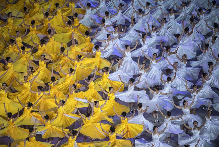 Hundreds of performers dance in formation together, wearing white or yellow flowing skirts.