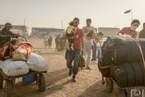 Photograph showing crowd of migrants walking with carts of their belongings; man holding small child