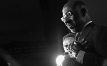 A black-and-white photo of Raphael Warnock wearing a suit and glasses in front of a spotlight, with his hands crossed over his heart