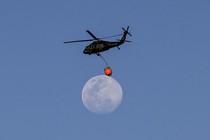 A firefighting helicopter carries a bucket as it flies past a nearly full moon.