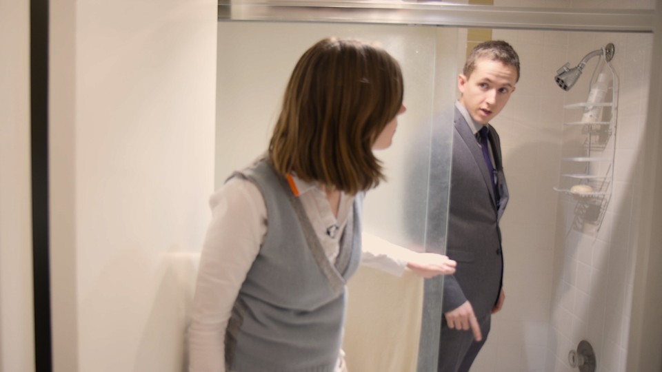 A woman peeking in on James Hamblin, who is fully dressed in a suit, in the shower