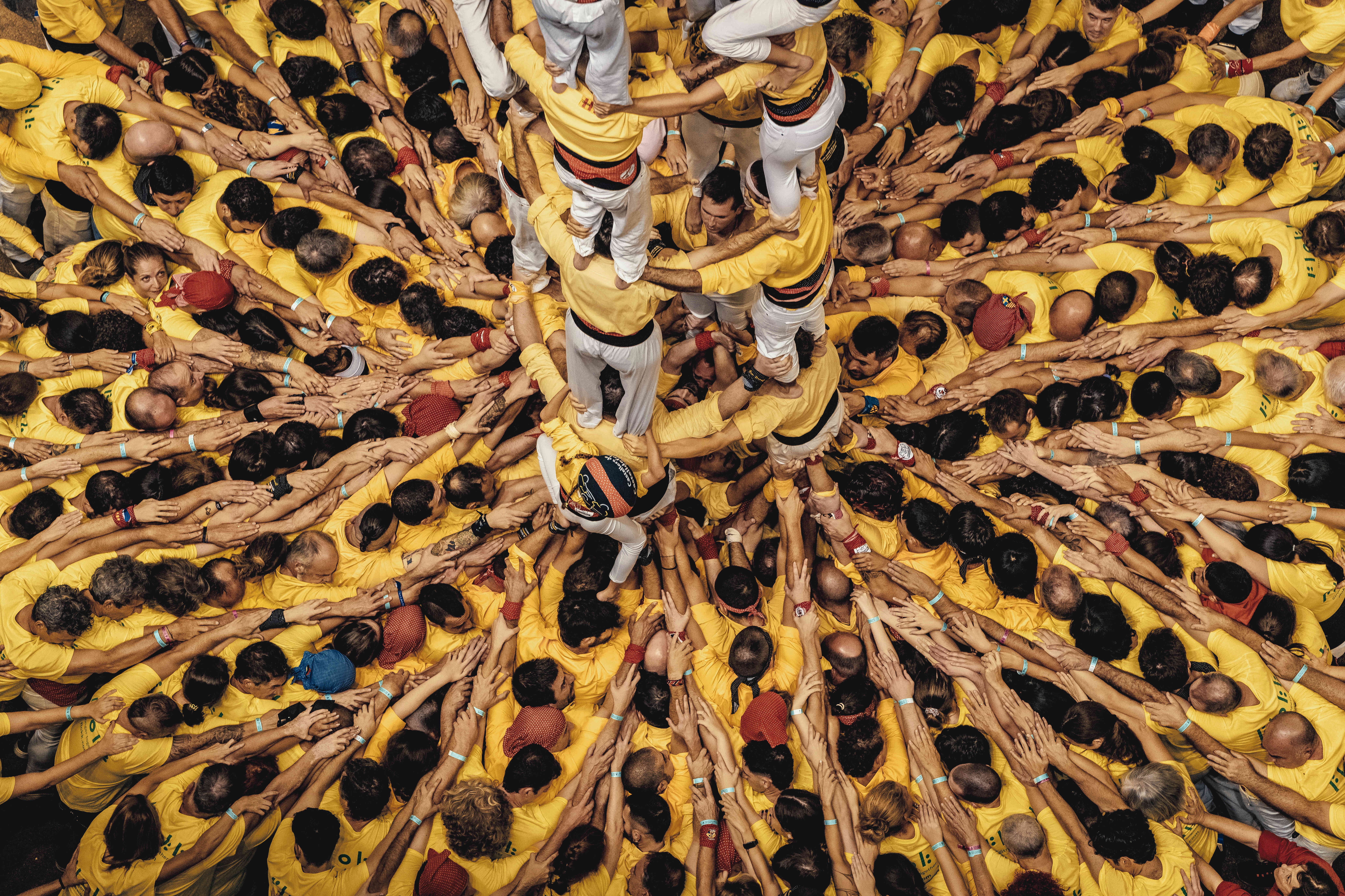 An overhead view of a large crowd of people wearing yellow, pusing inwards, arms linked over each other's shoulders, supporting a human tower in the center