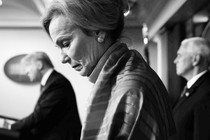 A black-and-white photo of Deborah Birx in the foreground, looking down, as Donald Trump stands at a lectern in the blurry background