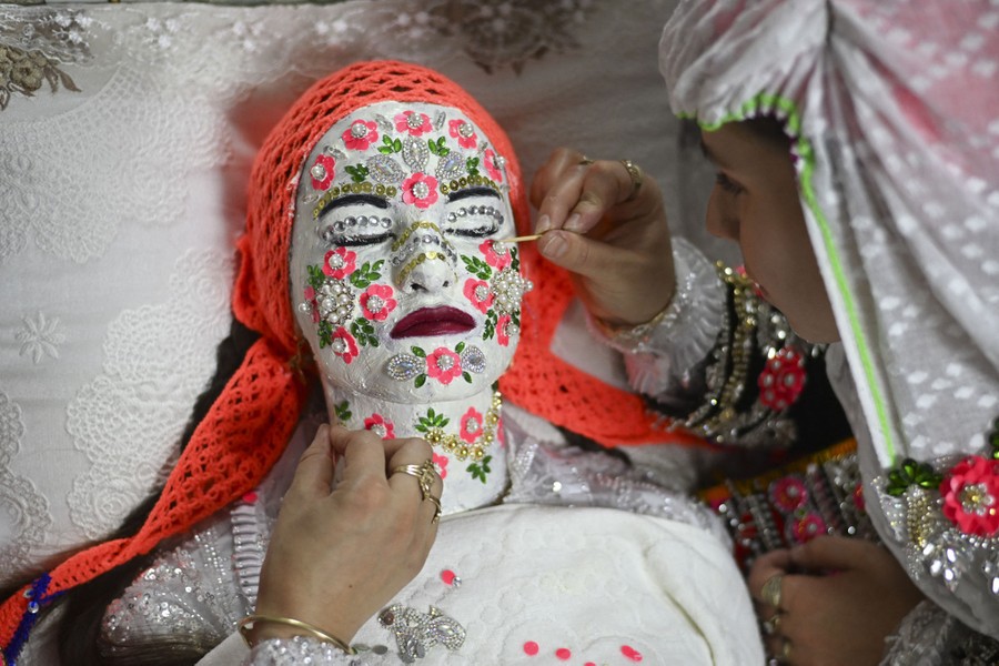 A woman lies still as others apply heavy colorful makeup all over her face and neck.