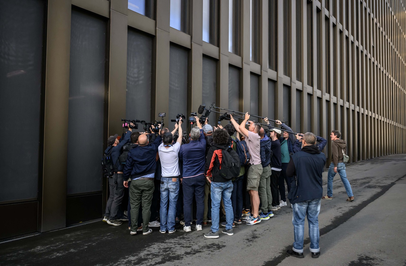 A tight group of journalists surround a lawyer, crowded against the outside wall of a building.