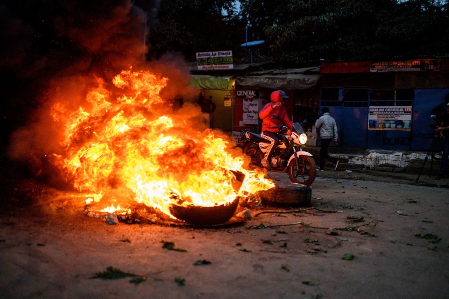 A person rides a motorcycle past a pile of burning tires in a street.