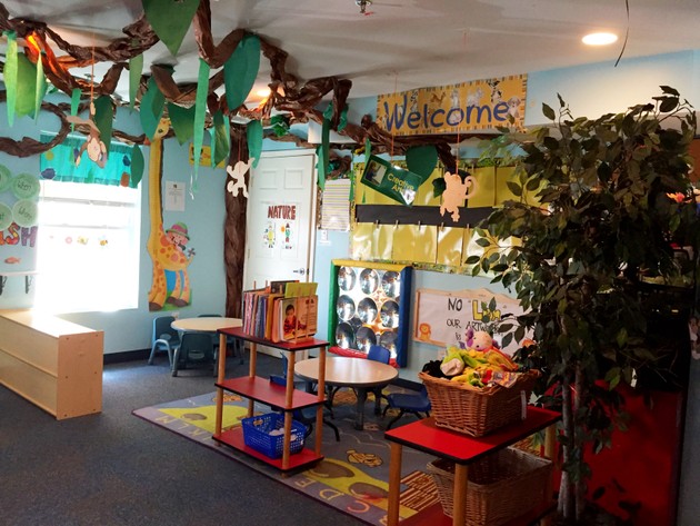 A colorful classroom with paper leaves and vines hanging from the ceiling and red tables with books and toys on the floor.