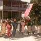 A photograph of group of Black people dressed in colorful summer celebration clothes in front of a hanging American flag and a two-story building