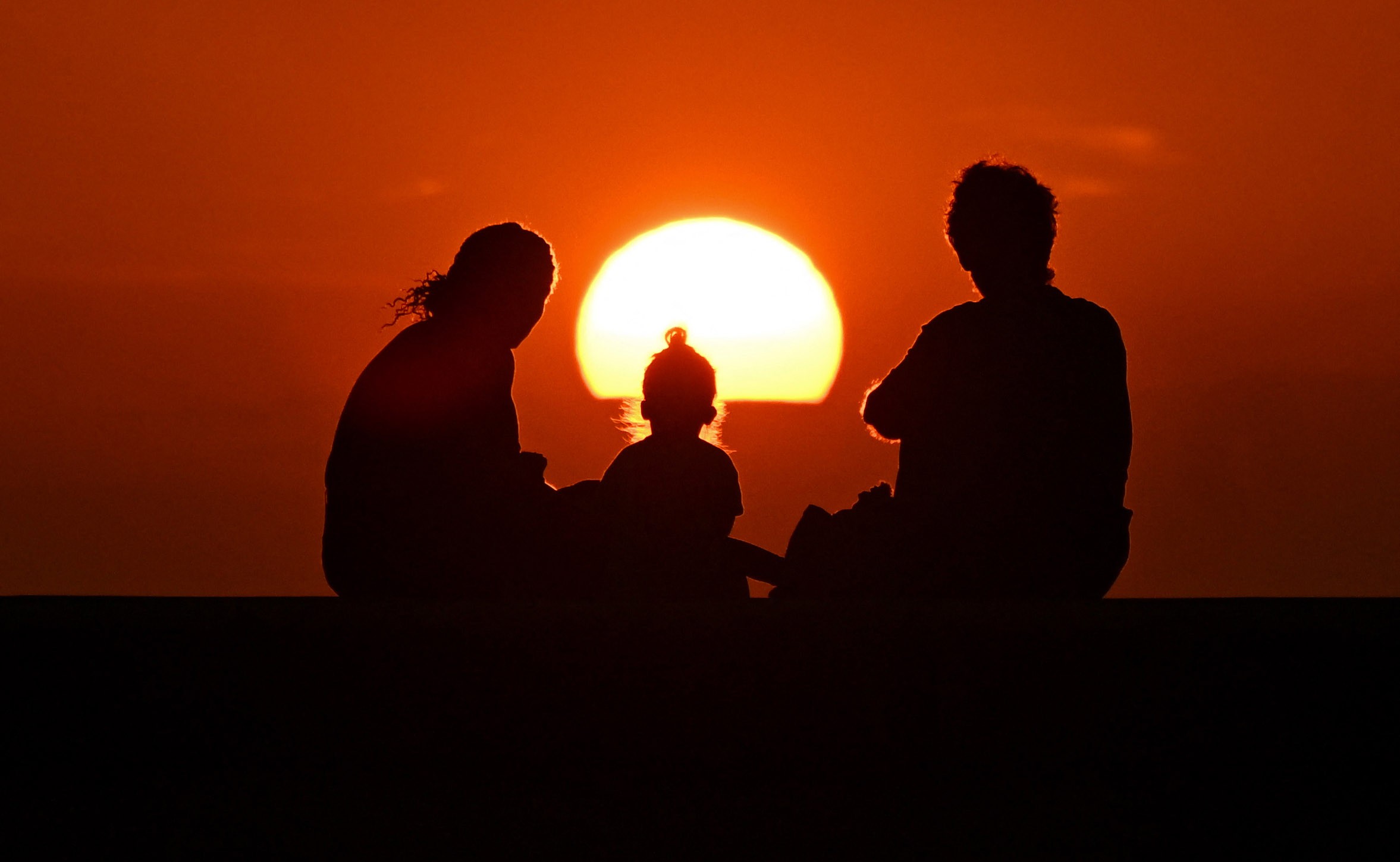 A family of three watches a sunset, seen in silhouette.