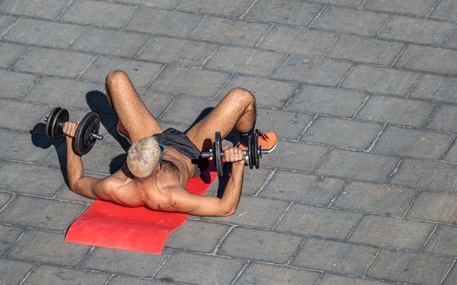 A shirtless man laying on a yoga mat and lifting weights.