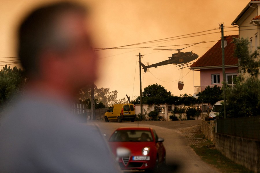 A firefighting helicopter carrying a water bucket flies low in a neighborhood.