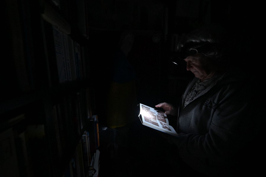 A person stands in front of a bookshelf in the dark, reading a book lit  by their headlamp.
