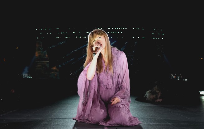 Taylor Swift kneeling in a lavender flowy robe holding a microphone