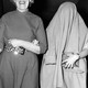 Black and white image of two women walking arm in arm, one with a big smile on her face, the other with a coat over her head, obscuring her face